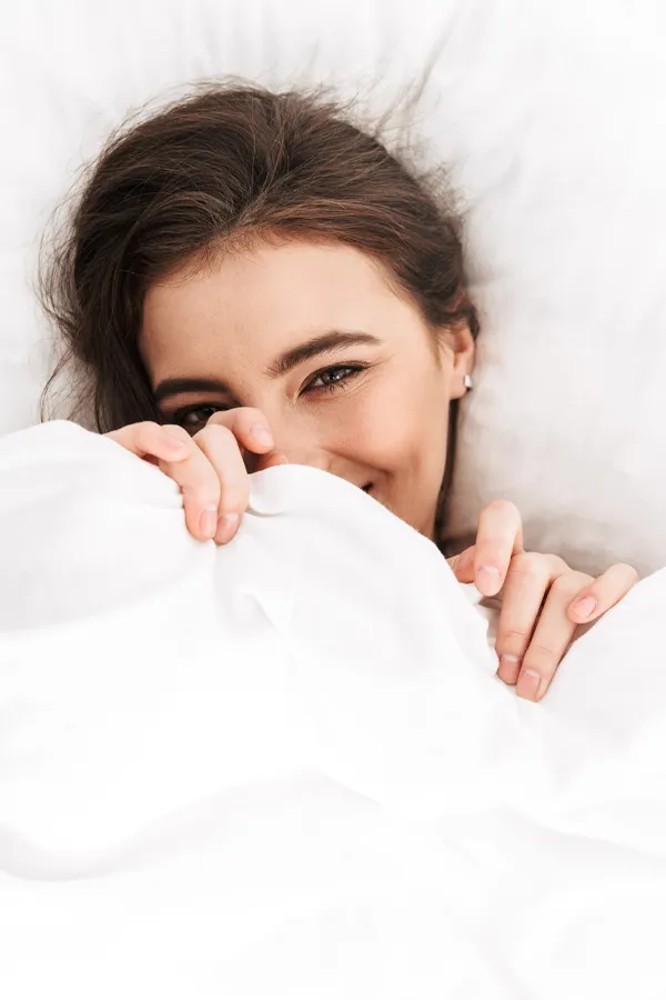 Apex-and-Intensity-Treatment A woman with long brown hair giggles in bed and pulls a sheet up over her face. Get Apex and Intensity treatment for vaginal health and stimulation from Regenerative Medical Institute in Greenville.