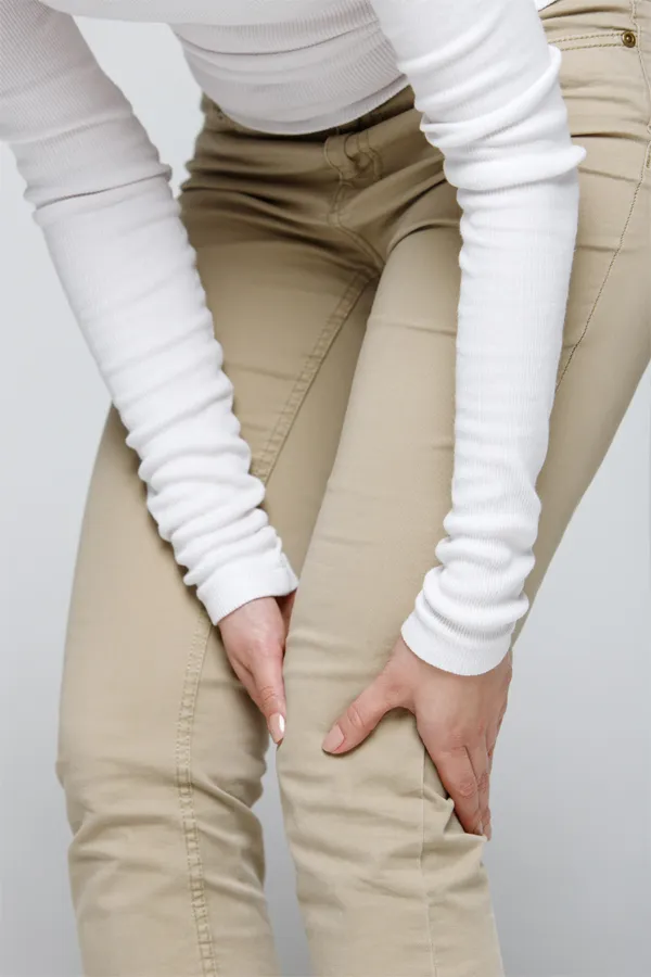 Arthritis-Treatment A woman wearing a white long-sleeve shirt and tan pants, clutching her knee, suffering from arthritis. Get arthritis treatment from Regenerative Medical Institute in Greenville.
