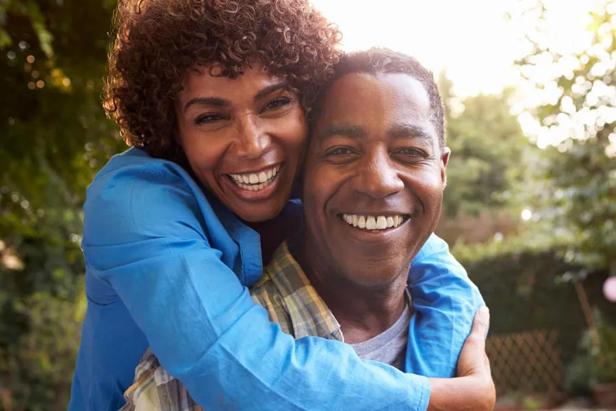 Autoimmune-Doctor A middle-aged black couple outside, the woman embracing the man from behind pressing her cheek to his, both are smiling due to successful Autoimmune Disorder treatment from Regenerative Medical Institute in Greenville.
