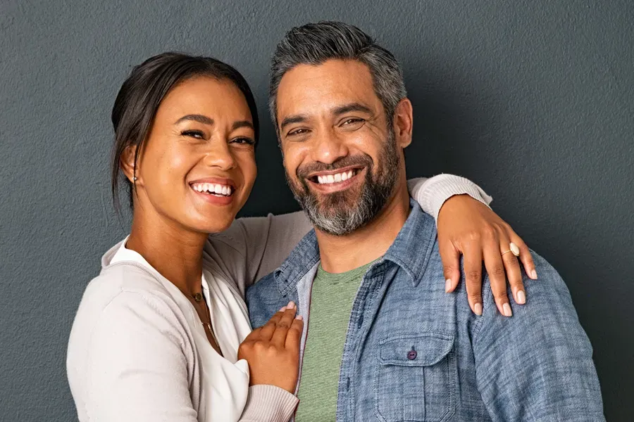 A middle-aged couple with their arms around each other standing against a gray background and smiling, happy with the bioidentical hormone therapy they received from Regenerative Medical Institute in Greenville.