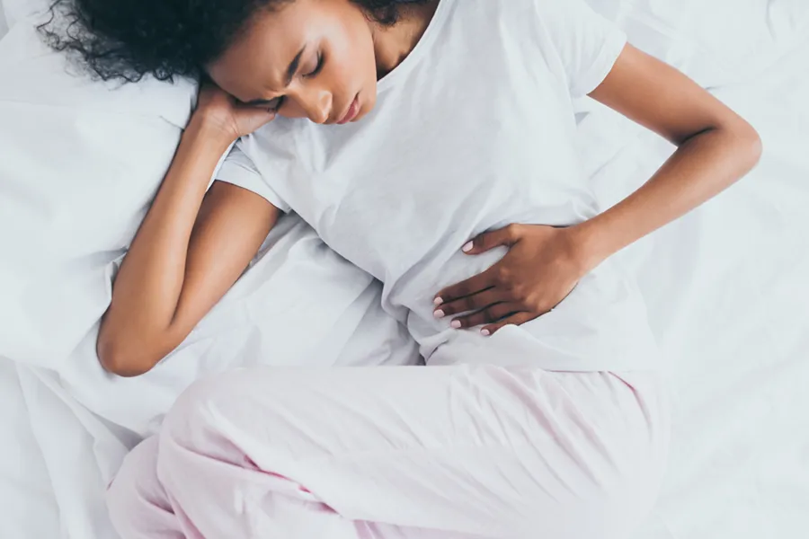 Crohns-Disease-Clinic A dark-skinned woman in white clothes lays on her bed clutching her middle before getting treatment for Crohn's Disease from Regenerative Medical Institute in Greenville.