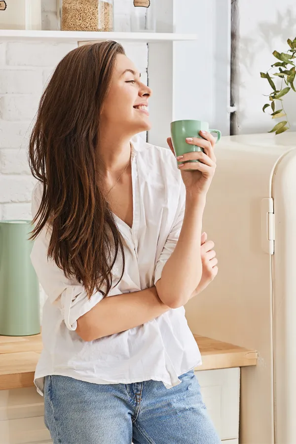 Crohns-Disease-Treatment A woman with a white blouse an jeans drinking tea in her kitchen, smiling after getting treatment for Crohn's Disease from Regenerative Medical Institute in Greenville.