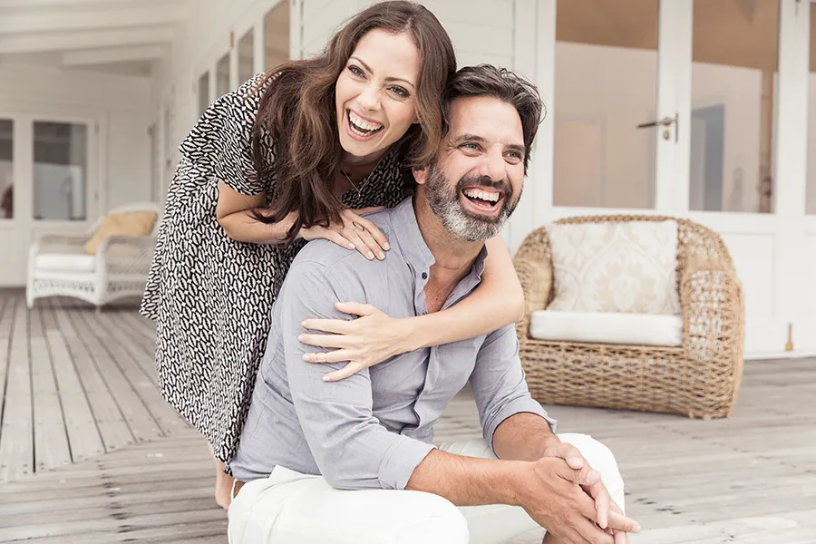 A middle-aged couple on a wooden porch with wicker furniture; the woman is playfully hugging the man from behind. They are benefiting from the health and wellness services from Regenerative Medical Institute in Greenville.