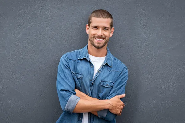 Hormones-for-Men-Doctor A man in a denim shirt stands smiling against a gray-blue wall, pleased with his testosterone hormone therapy from Regenerative Medical Institute in Greenville.