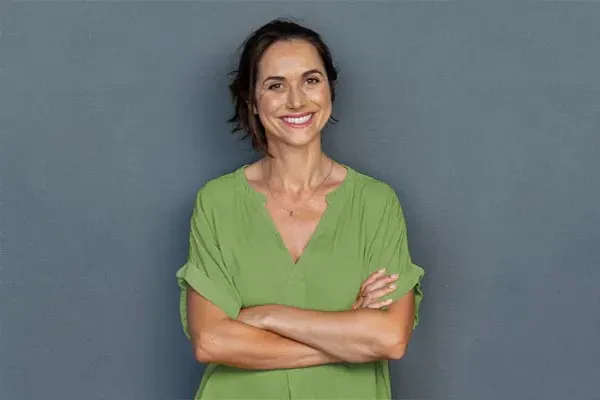 Hormones-for-Women-Doctor A woman in a green shirt stands smiling against a gray-blue wall, pleased with her hormone therapy from Regenerative Medical Institute in Greenville.