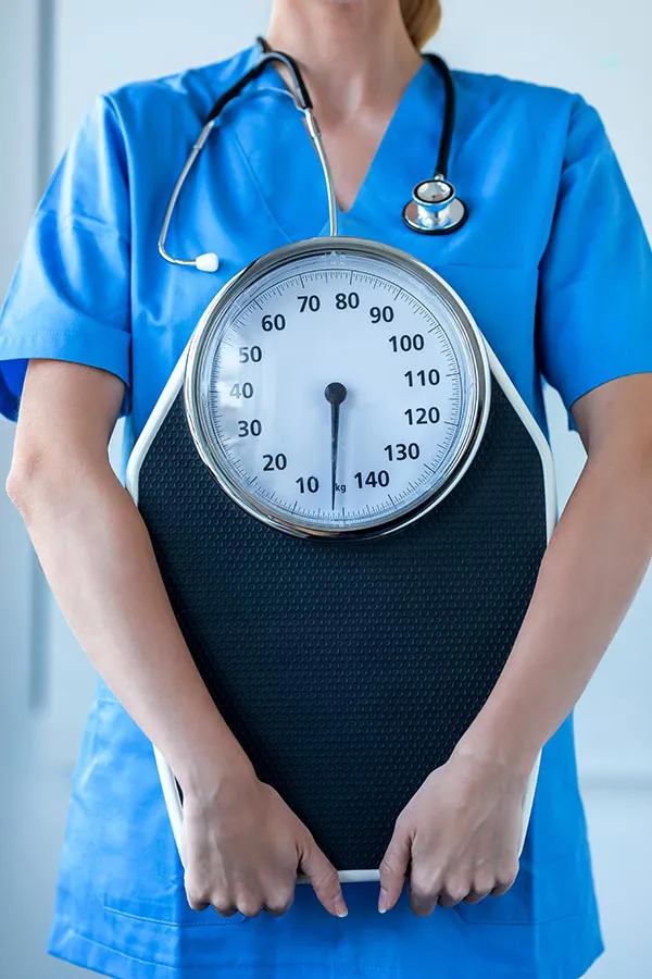 Medical-Weight-Loss-Treatment A nurse in blue scrubs with a stethoscope hanging around her neck holds a scale, representing weight loss treatments from Regenerative Medical Institute in Greenville.