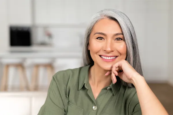 Menopause-Clinic An asian woman with long gray hair and a green shirt sitting and smiling on her couch, happy with her menopause care from [NAME} in Greenville.