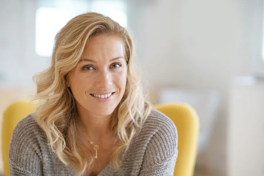 Perimenopause-Doctor A blonde woman wearing a a gray sweater sits inside in a yellow chair, smiling and happy with her perimenopause treatment from Regenerative Medical Institute in Greenville.