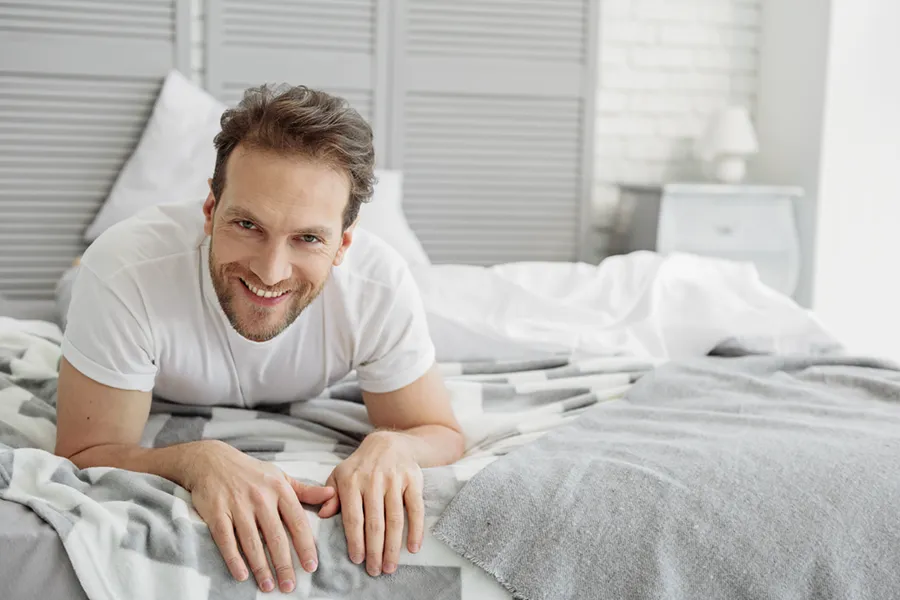 Sexual-Wellness-for-Men-Doctor A man in a t-shirt lies on his stomach in bed smiling, happy with his sexual wellness treatments from Regenerative Medical Institute in Greenville.