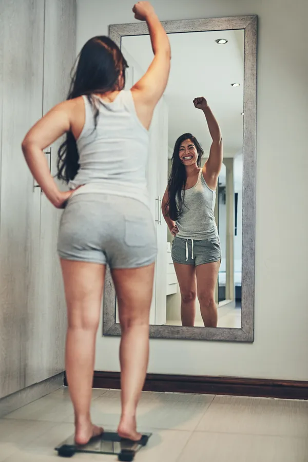 Tirzepatide-Treatment A woman in workout clothes stands on a scale looking in the mirror and pumping her hands in celebration of her successful Tirzepatide treatment from Regenerative Medical Institute in Greenville.