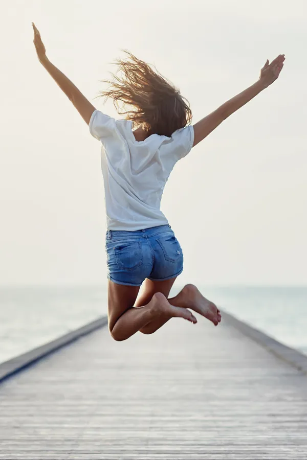 Votiva-Treatment A woman in denim shorts and a white t-shirt jumps for joy on a pier. Get Votiva vaginal rejuvenation treatment from Regenerative Medical Institute in Greenville.